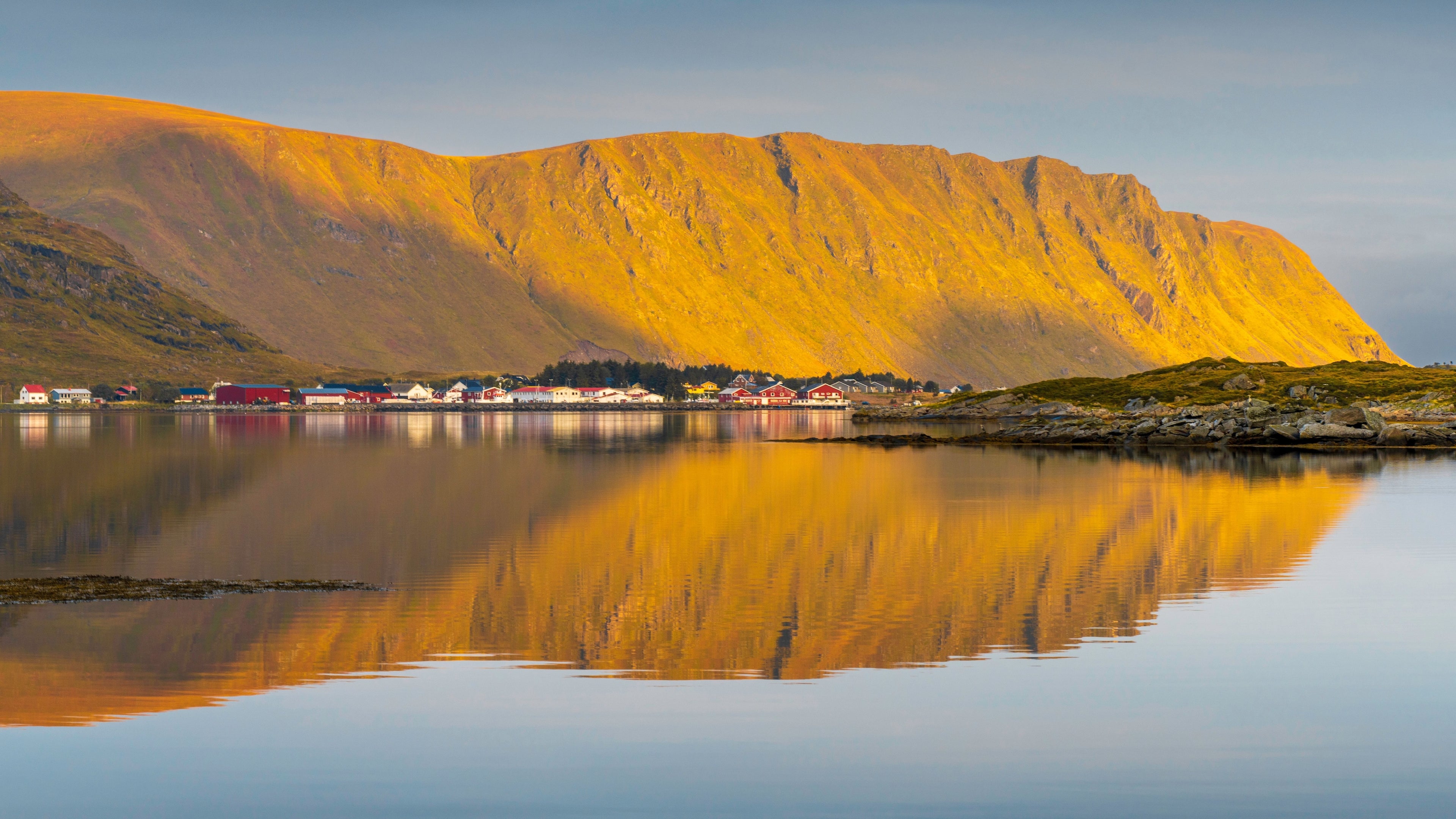 Fotografie eines Fjordes im Sommer mit einfallendem Sonnenlicht bei Sonnenuntergang. Im Hintergrund befindet sich eine Siedlung vor einem hohen Berg.